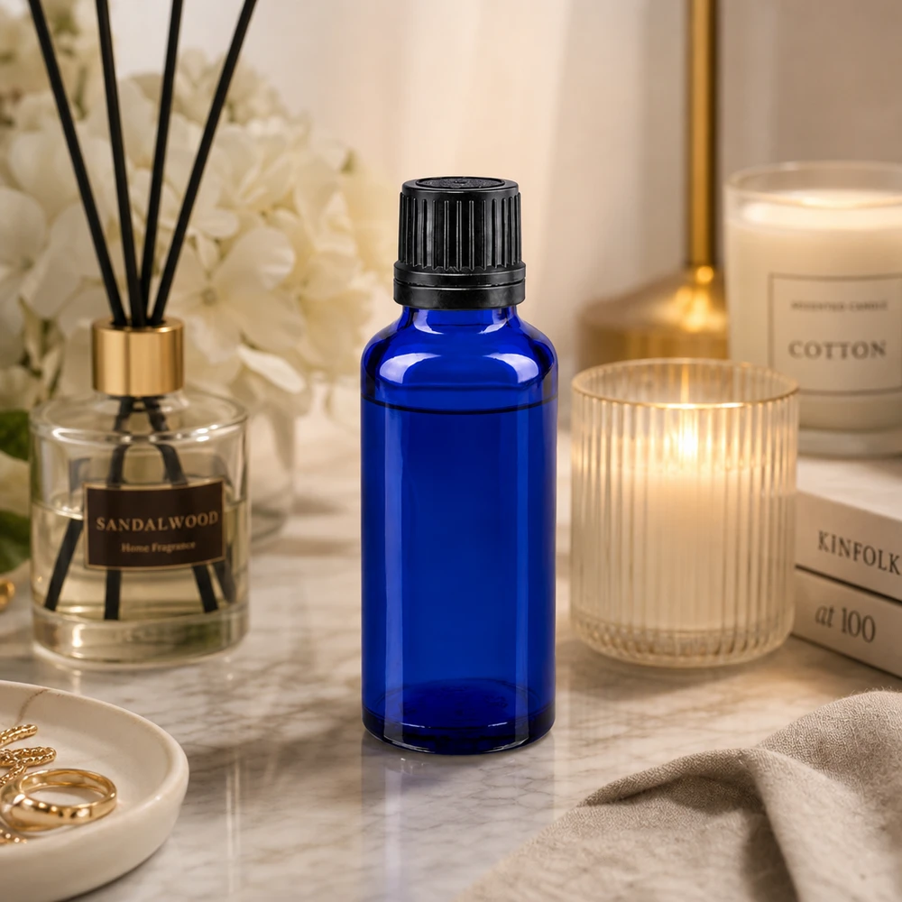 Apoteksflaske blå 30ml skruelåg sort hældeprop r. OV Blå 30 ml glasflaske med sort dråbepropp på marmor ved siden af et duftlys og en rumduft