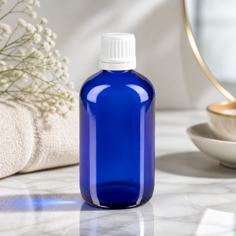 Blå glasflaske på 100 ml med hvid OV-lukning på marmor ved siden af et håndklæde og tørrede blomster