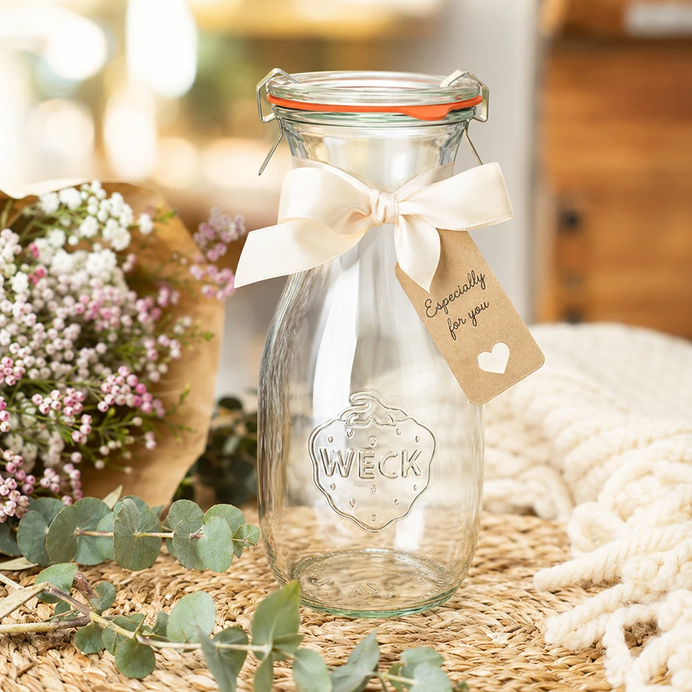 530 ml saftflaske, komplet WECK RR60 WECK 530 ml juiceflaske RR60 dekoreret med et hvidt bånd og et gavemærke arrangeret mellem blomster