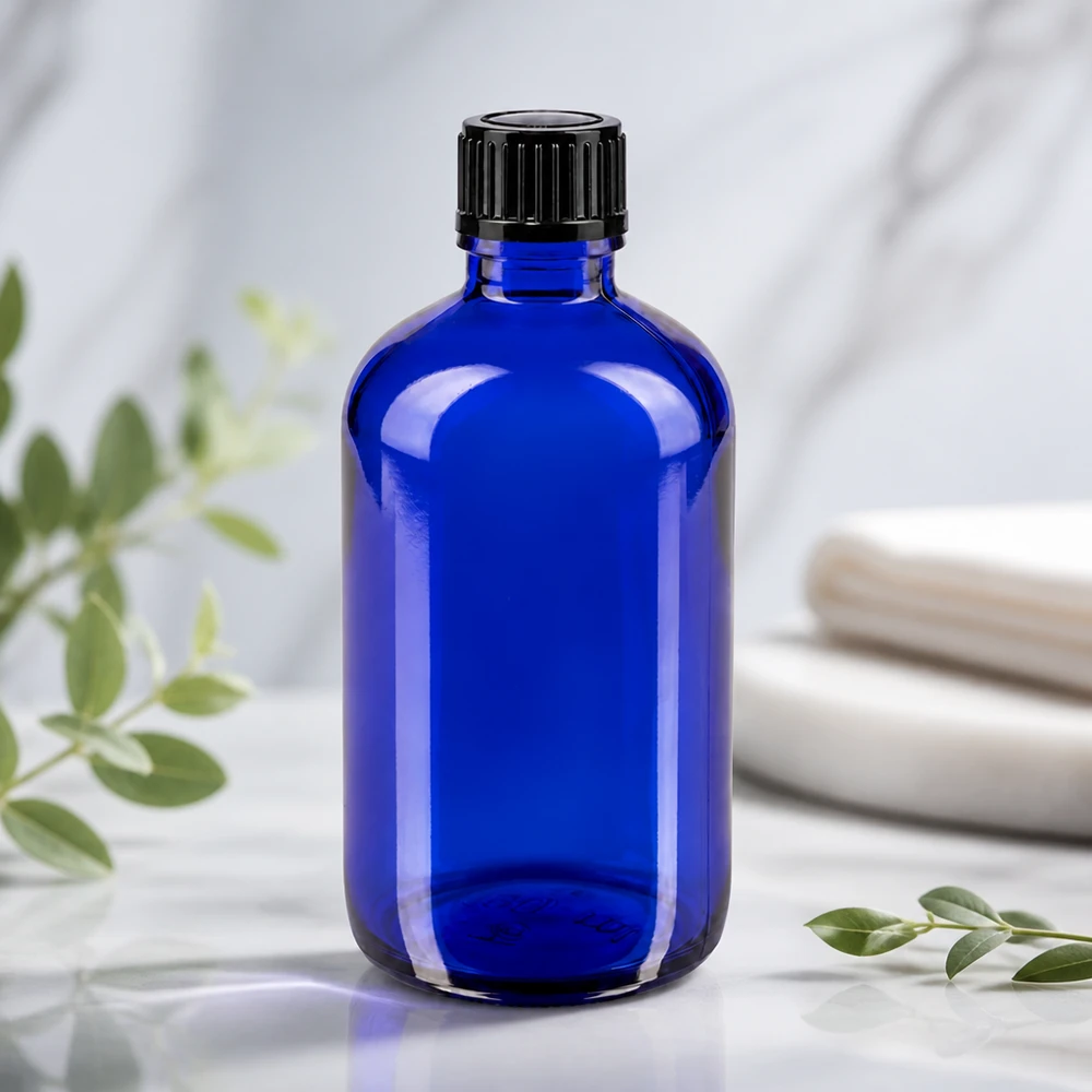 Apoteksflaske blå 100ml dryp v. 1mm sort pc Blå glasflaske på 100 ml med riflet sort prop på en hvid marmoroverflade med eukalyptusgrene