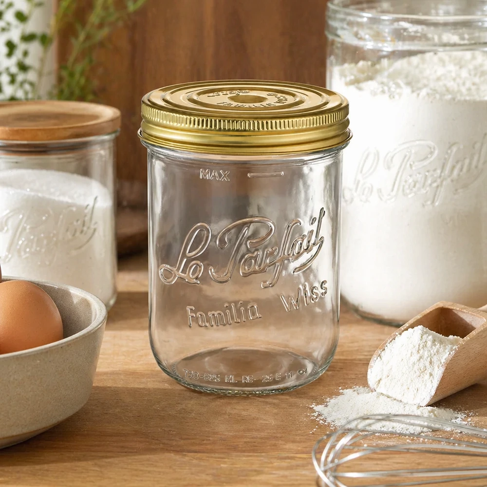 Le Parfait® Familia Wiss 1 kg Ø100 mm glas 1020 ml Familia Wiss-krukke med gyldent låg på træbord ved siden af bageingredienser og andre konserveringskrukker