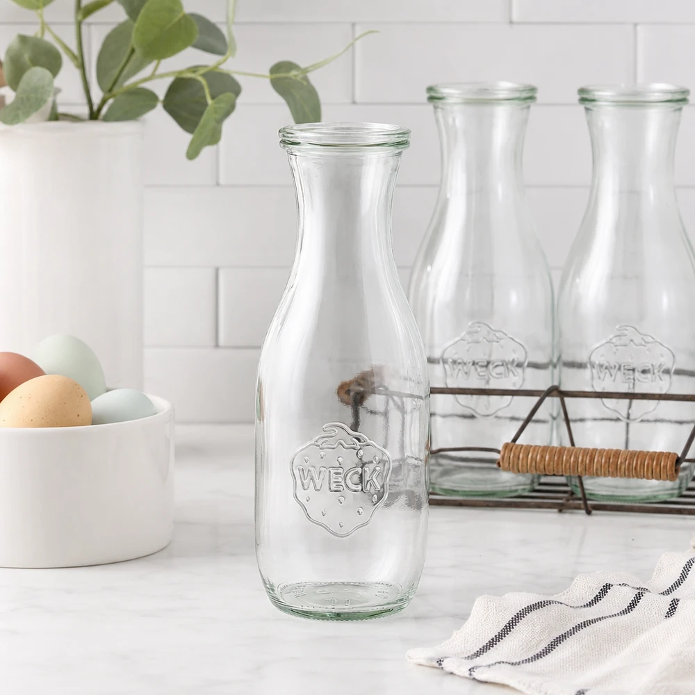 1062 ml saftflaske med glaslåg WECK RR60 WECK juiceflaske 1062ml glasflaske tom i moderne hvidt køkken med æggeskalstrådkurv og plante