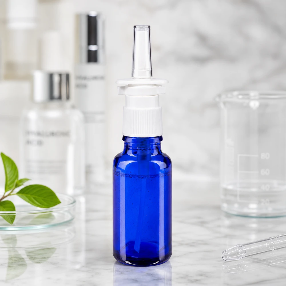 Næsesprayflaske på 20 ml af blåt apoteksglas med hvid forstøver ved siden af et bægerglas og hyaluronsyre