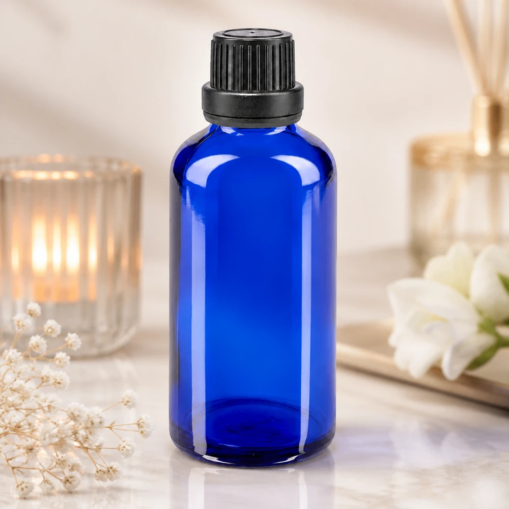 Koboltblå glasflaske på 50 ml med sort skruelåg og dråbeindsats ved siden af et lys og blomster