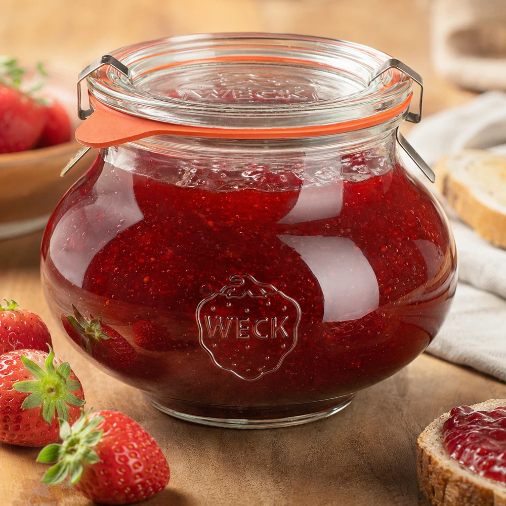 560 ml dekorativt glas, komplet WECK RR80 WECK dekorativ krukke 560 ml fyldt med hjemmelavet jordbærmarmelade, lukket svinglåg, omgivet af frisk frugt og grøntsager.